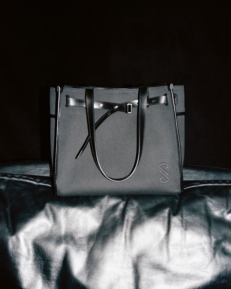 Elegant gray canvas tote bag with black leather trim and handles displayed on a reflective silver surface against a dark backdrop.