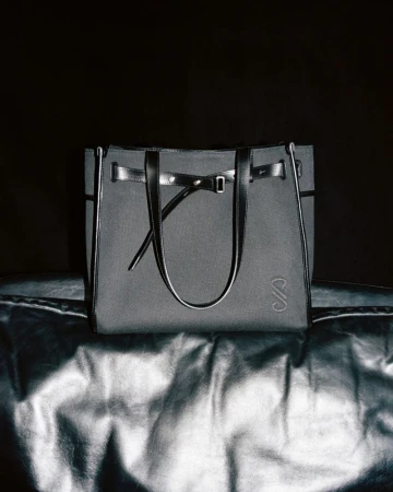 Elegant gray canvas tote bag with black leather trim and handles displayed on a reflective silver surface against a dark backdrop.