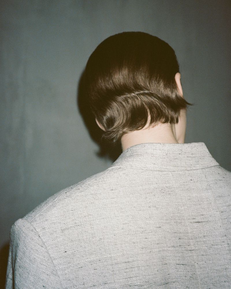 Woman with dark, curled bob hairstyle, wearing a textured grey jacket, posing in front of a grey backdrop in a fashion studio.
