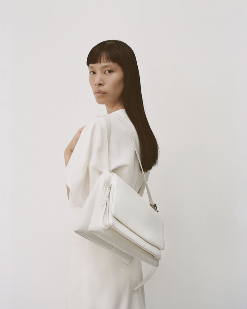 Studio shot of a slender Asian woman with straight black hair, wearing an off-white dress and carrying a white leather handbag.