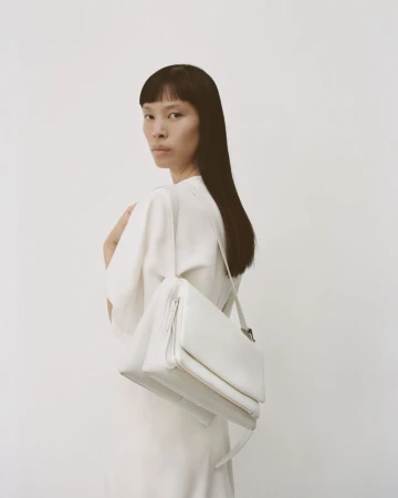 Studio shot of a slender Asian woman with straight black hair, wearing an off-white dress and carrying a white leather handbag.