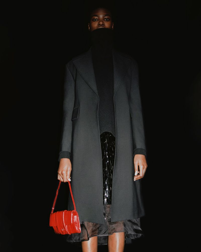 Fashionable woman in a dark coat and turtleneck holding a red purse against a dark backdrop in a studio fashion shot.
