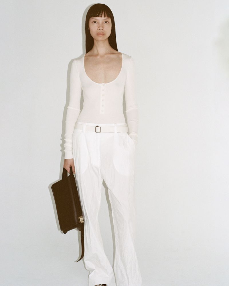 Elegant Asian woman posing in long-sleeve top and white pants, holding a structured handbag against a bright white backdrop.