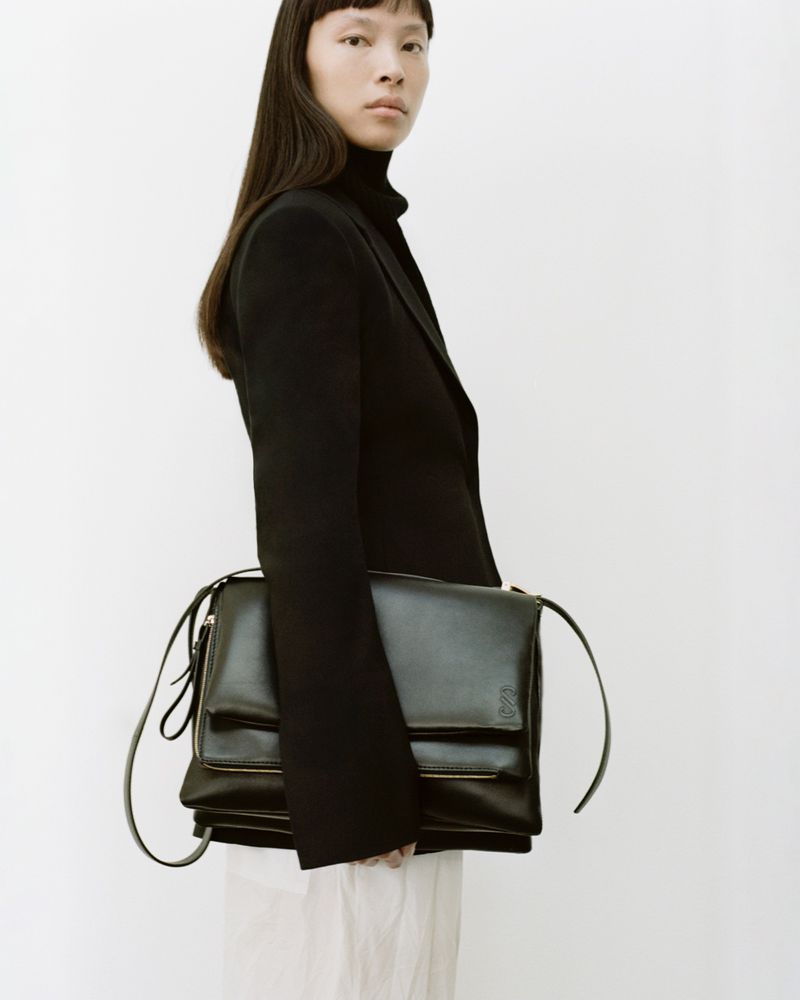 Asian fashion model poses in a studio wearing a black turtleneck, jacket, and carrying a black leather handbag against a white backdrop.