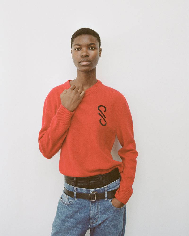 Young woman poses in red sweater and jeans against white wall in a fashion shot with natural light. Neutral expression.