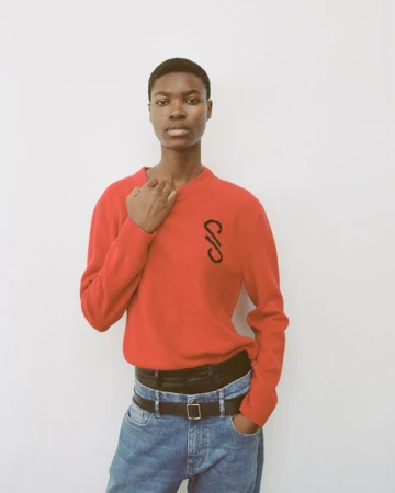 Young woman poses in red sweater and jeans against white wall in a fashion shot with natural light. Neutral expression.