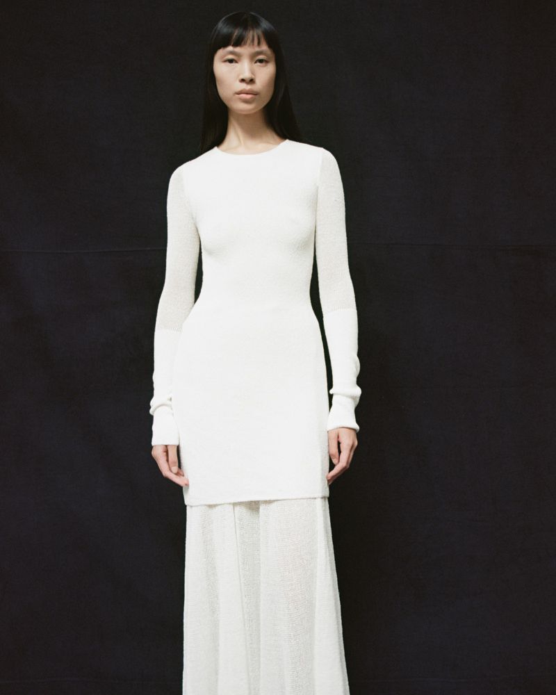 Elegant woman in white long-sleeve sweater dress and skirt against black backdrop, fashion editorial, indoor studio shot.