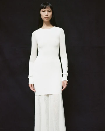 Elegant woman in white long-sleeve sweater dress and skirt against black backdrop, fashion editorial, indoor studio shot.