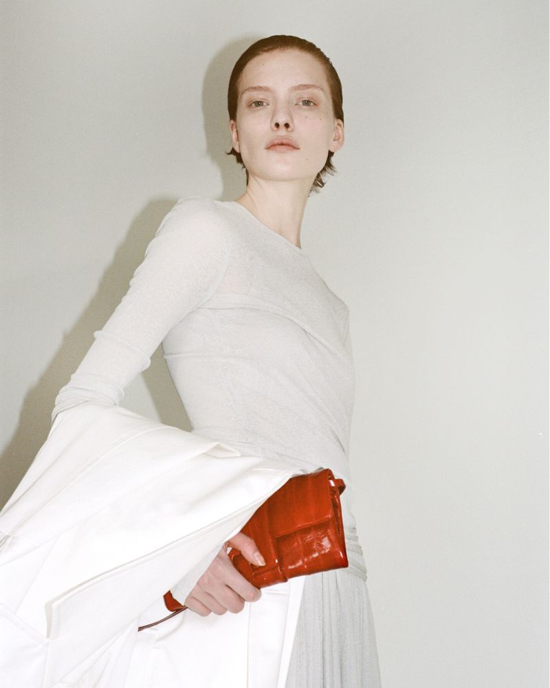 Woman in white poses with red handbag in studio. Minimal fashion shot with soft lighting and neutral background.