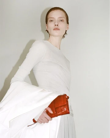 Woman in white poses with red handbag in studio. Minimal fashion shot with soft lighting and neutral background.