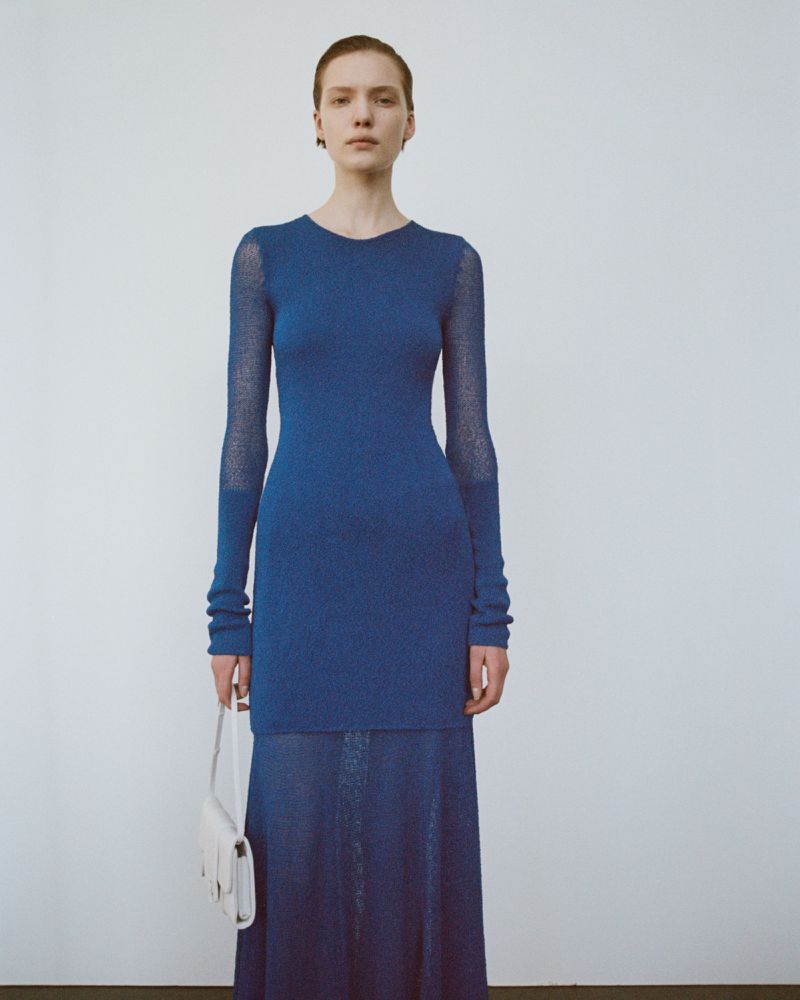 An adult female fashion model poses in a long-sleeved blue knit dress with a white purse in a studio setting with a neutral background.
