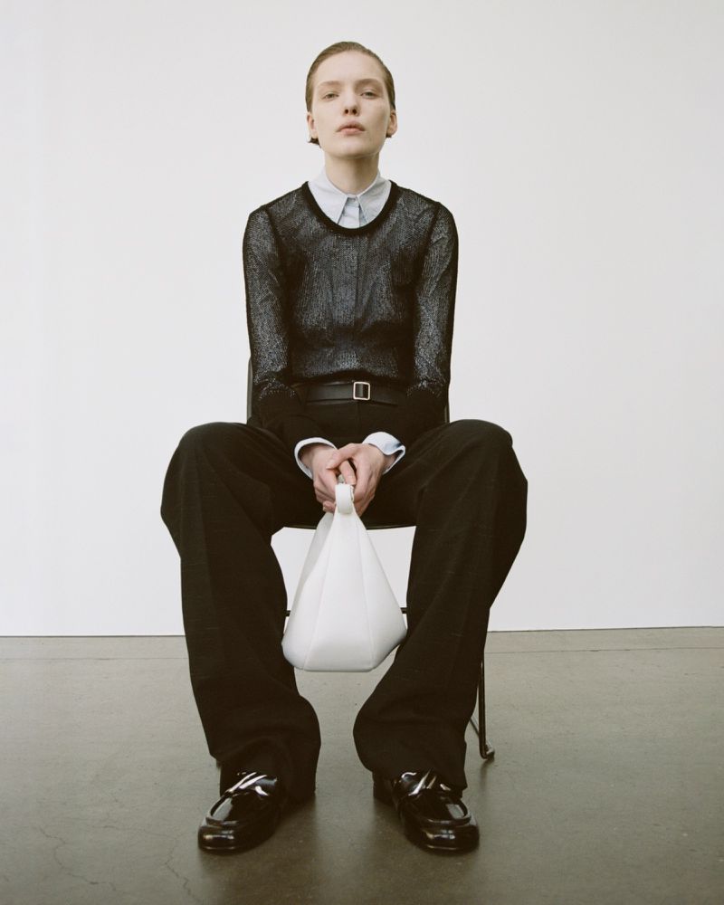 A person is sitting on a chair holding a white bag, with black pants and Mary Jane shoes against a neutral backdrop in a fashion shot.
