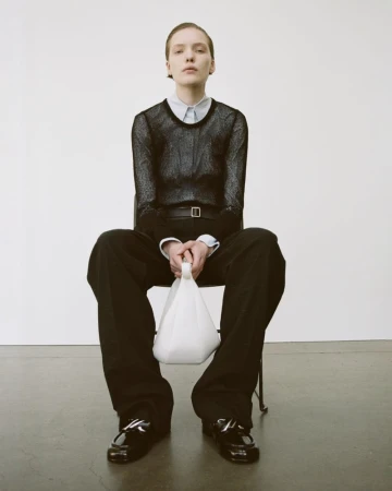 A person is sitting on a chair holding a white bag, with black pants and Mary Jane shoes against a neutral backdrop in a fashion shot.