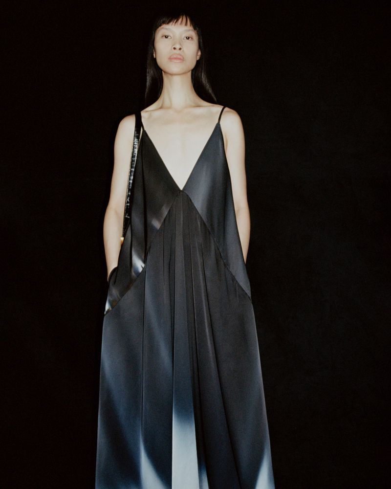 Asian woman stands in a long black dress with spaghetti straps against a dark background in a studio fashion photograph.
