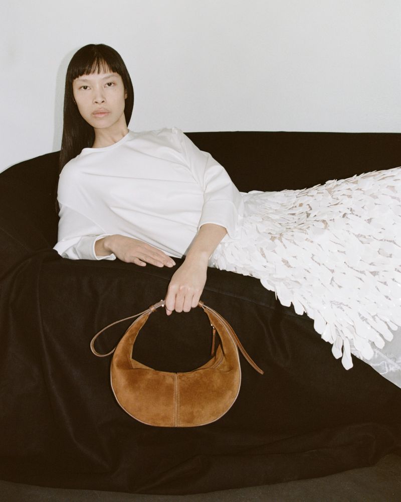 Adult woman posing on a black couch with a brown purse and wearing a white dress in a studio setting with soft lighting.