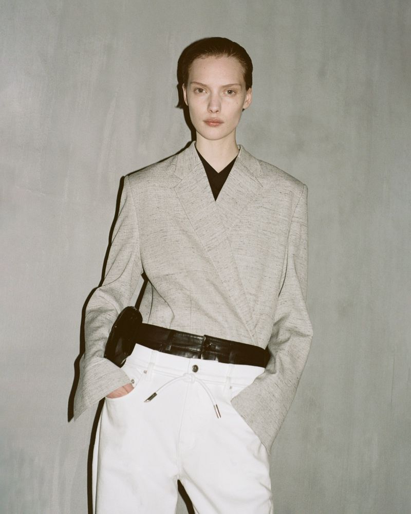 Fashionable adult woman poses indoors wearing a light grey blazer, black top, white pants, and black belt in a fashion editorial.
