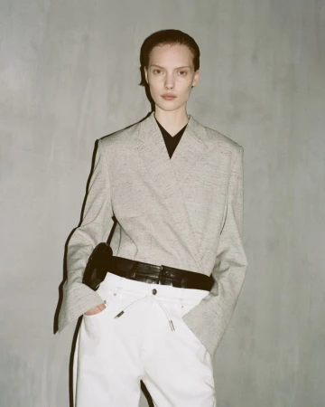 Fashionable adult woman poses indoors wearing a light grey blazer, black top, white pants, and black belt in a fashion editorial.