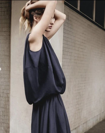 Adult female fashion model in a sleeveless, dark blue dress with a brick wall background, posing with hands behind her head.