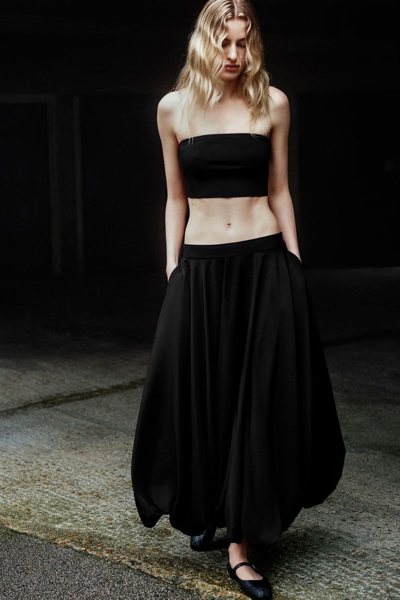 Young woman in a black tube top and long skirt posing against a dark background, showcasing fashion and minimalist style with soft, diffused lighting.