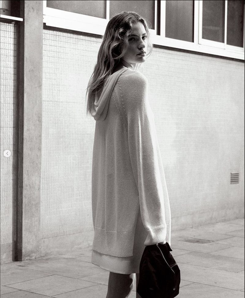 Blach-and-white shot of a female model with long hair posing in hooded sweater, standing outdoors against a concrete wall background.