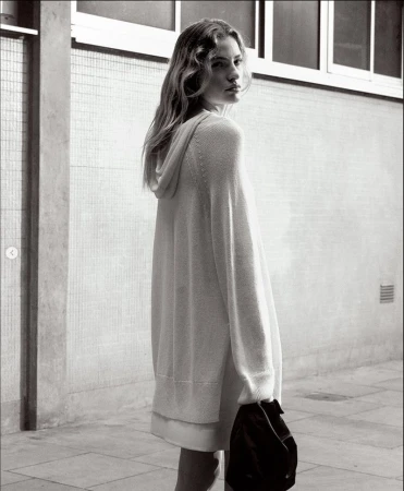 Blach-and-white shot of a female model with long hair posing in hooded sweater, standing outdoors against a concrete wall background.
