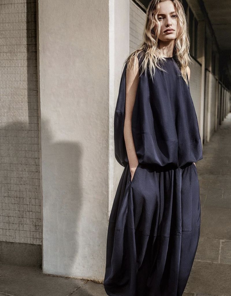 A fair-skinned woman with blonde hair poses in a stylish, oversized navy blue dress in a neutral indoor setting, fashion photography.