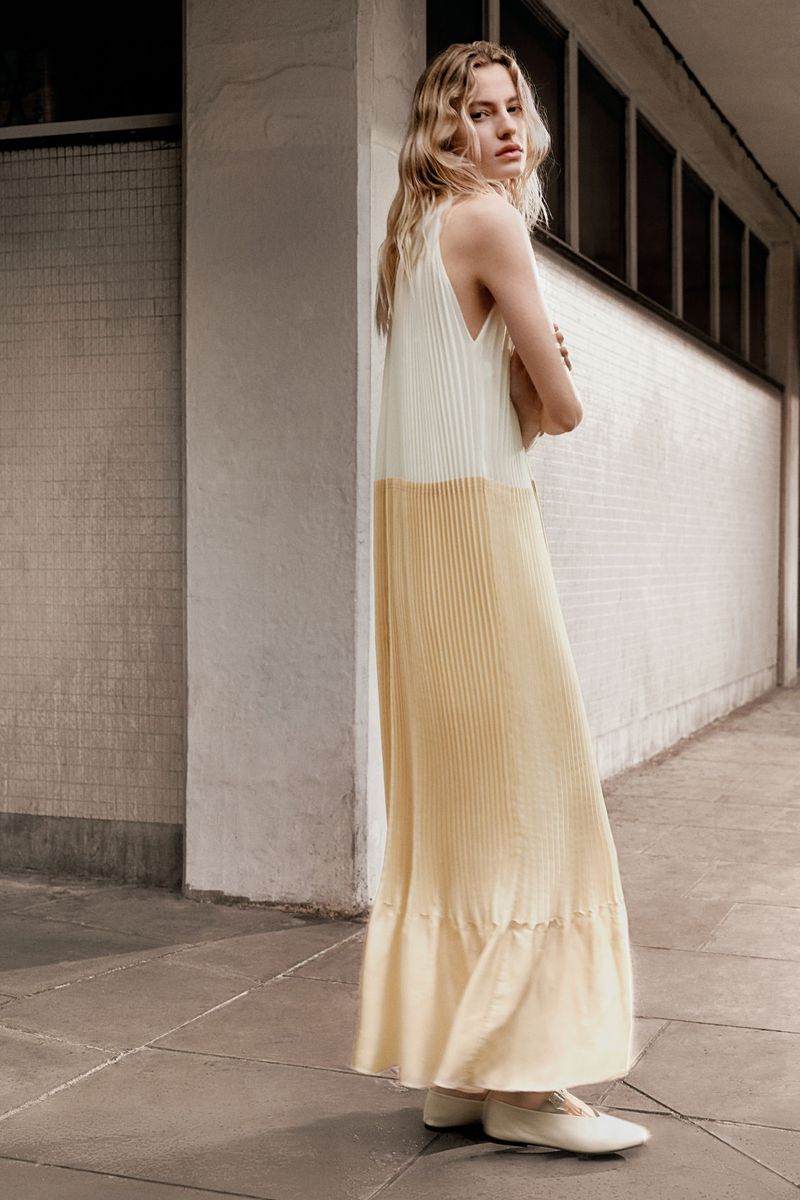 A blonde-haired adult female stands in a long, pleated white-and-yellow dress, wearing flats and posing in an urban outdoor setting.