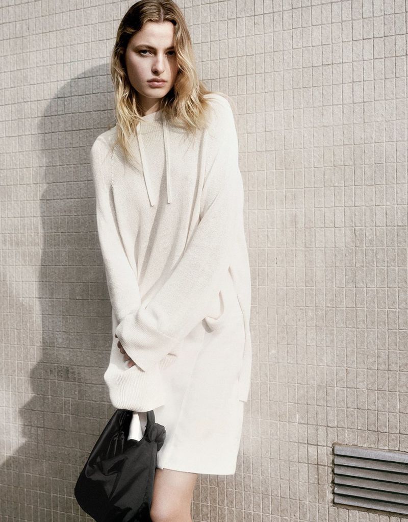 Fashion shot of a young adult woman in an off-white sweater dress, holding a black bag, posing against a beige tile wall indoors.