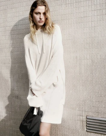 Fashion shot of a young adult woman in an off-white sweater dress, holding a black bag, posing against a beige tile wall indoors.
