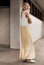Model wearing a long pleated cream and yellow dress standing outside against a concrete building wall