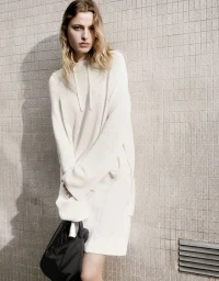 Model Felice Nova Noordhoff wearing a white knit dress and holding a dark handbag against a tiled wall