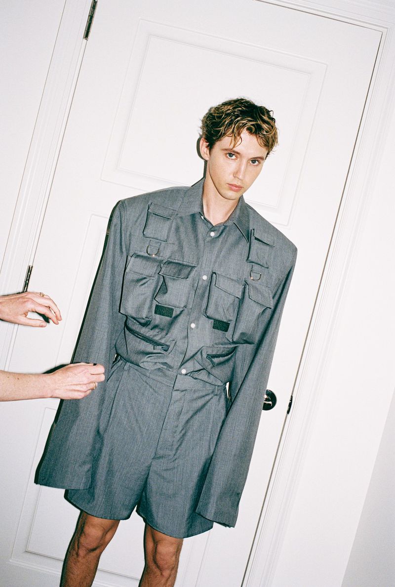 Young adult male in grey utility shirt and shorts stands in front of a white door, fashion shot, with a partially visible person.