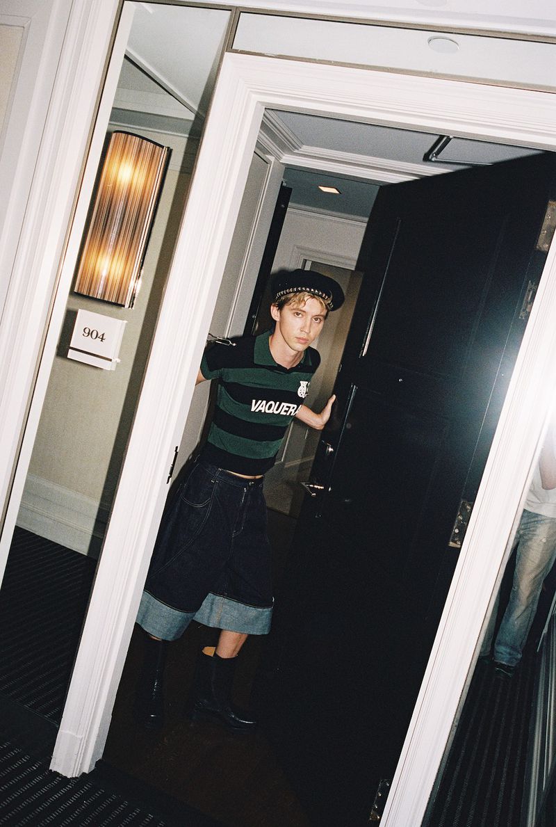 Stylish young man in a green and black striped shirt and denim shorts stands in a doorway inside a building with dark floors.