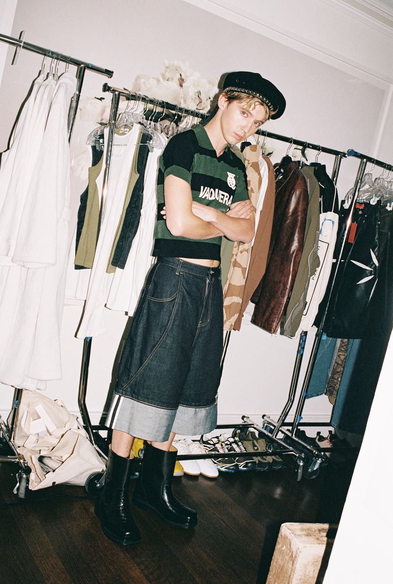 Young adult in black-and-green rugby shirt, denim culottes, and black boots poses indoors with racks of clothing.
