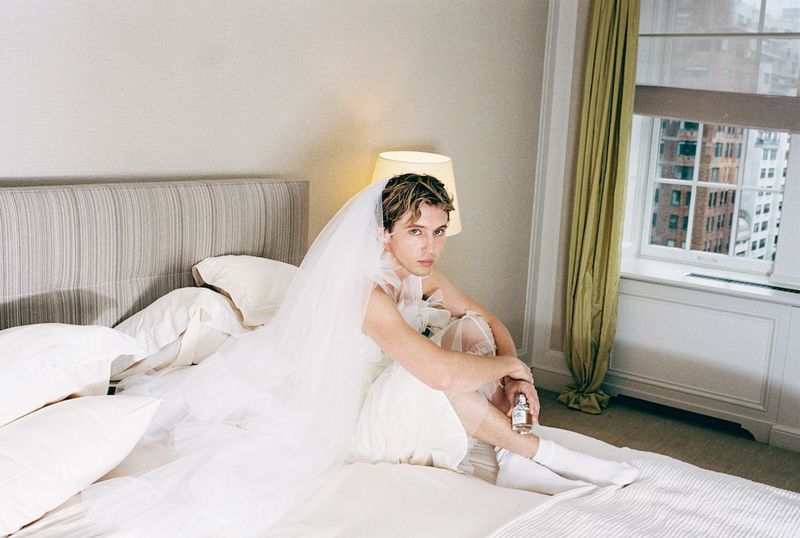 Male in a wedding dress and veil sitting on a bed in a room with a window and buildings in the background. 