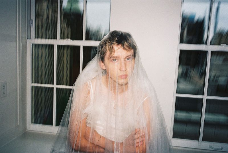 Portrait of an adult male indoors wearing a white top and draped with white tulle fabric posing in front of a window.