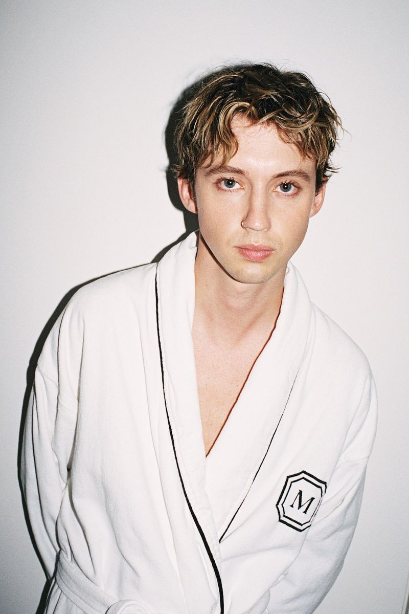 Portrait of an adult male wearing a white robe with blond hair standing in front of a white wall in a studio setting.