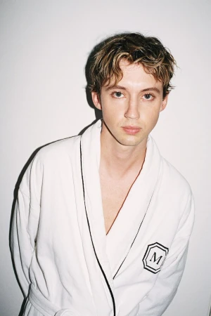 Portrait of an adult male wearing a white robe with blond hair standing in front of a white wall in a studio setting.