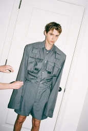 Troye Sivan wearing a structured grey utility shirt and shorts in front of a white panel door