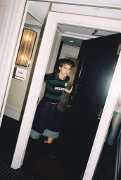 Troye Sivan wearing a striped Vaquera polo shirt, beret, and wide denim shorts posing in a hotel doorway.