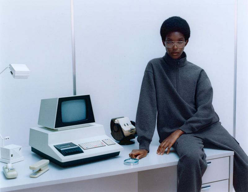 A person in grey sweatshirt and sweatpants sits on a desk with a vintage computer, tape drive, stapler, and desk lamp in a bright studio.