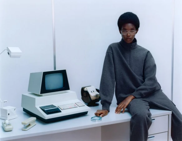 A person in grey sweatshirt and sweatpants sits on a desk with a vintage computer, tape drive, stapler, and desk lamp in a bright studio.