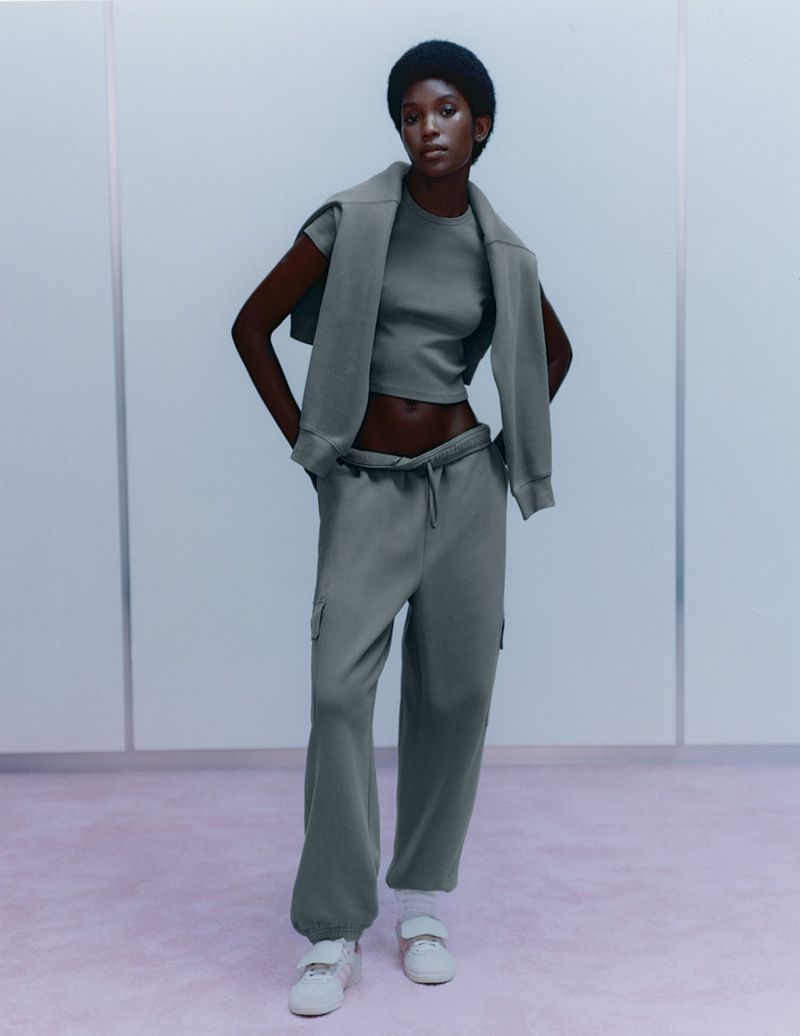 Fashion shot of Black female model in gray crop top, sweatshirt draped over shoulders, sweatpants, white sneakers against pale panel wall.