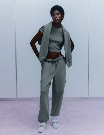 Fashion shot of Black female model in gray crop top, sweatshirt draped over shoulders, sweatpants, white sneakers against pale panel wall.
