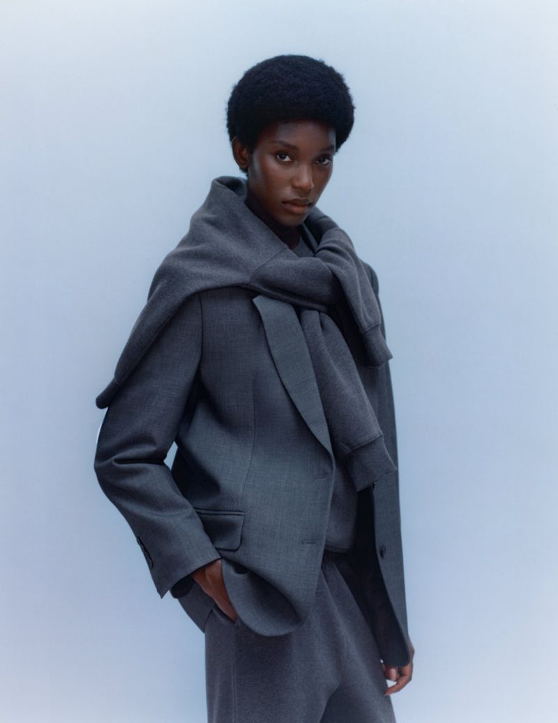Portrait of a slender Black female model in a grey suit with a sweater draped over her shoulders against a pale blue background.