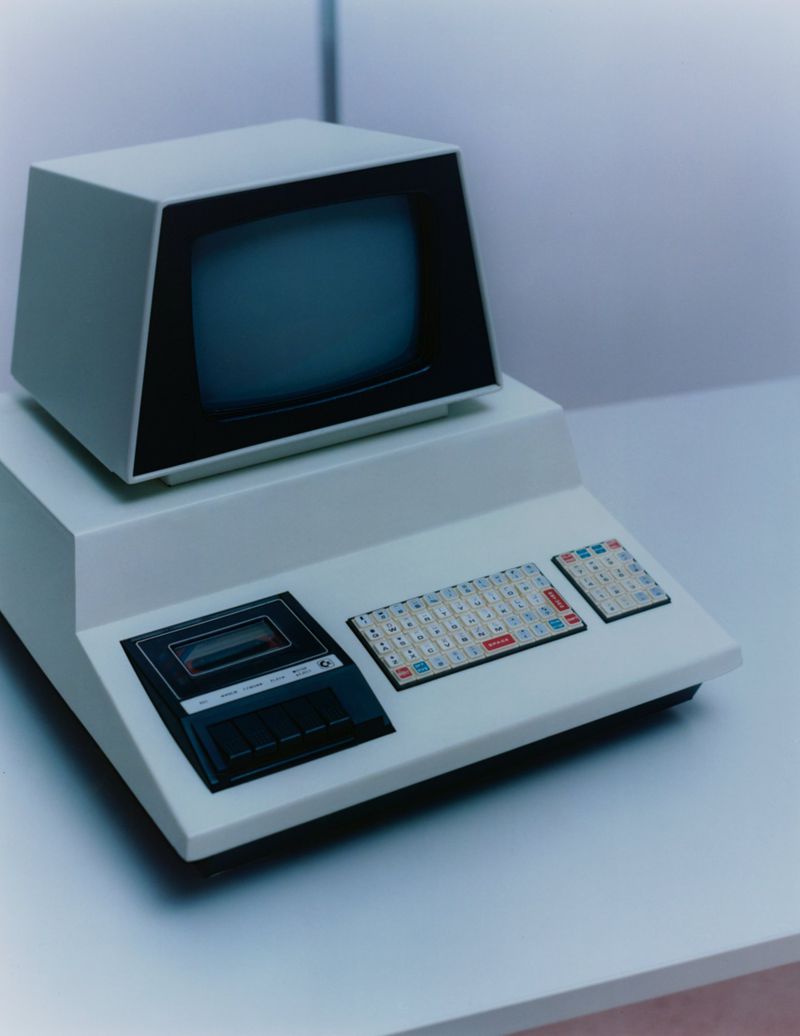 Vintage computer featuring a black screen and keyboard sits on a white desk against a plain wall in a softly lit indoor setting.
