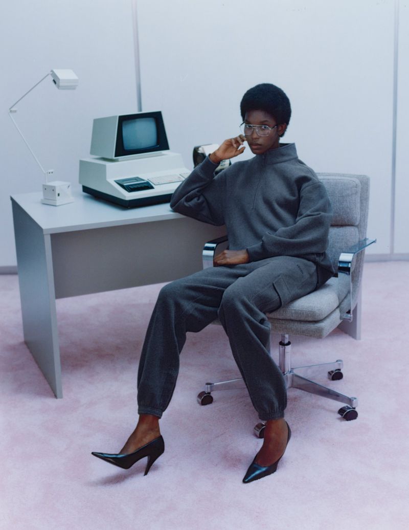 An adult woman with an afro and glasses sits at a desk with an old computer and lamp in a pale studio, wearing a grey sweatsuit and heels.
