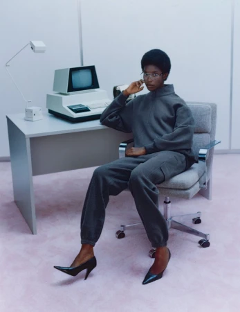 An adult woman with an afro and glasses sits at a desk with an old computer and lamp in a pale studio, wearing a grey sweatsuit and heels.