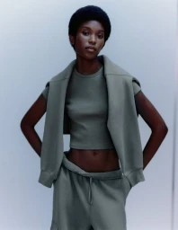 Model wearing olive green crewneck tee and sweatpants with a matching sweatshirt draped over shoulders against a grey studio backdrop.