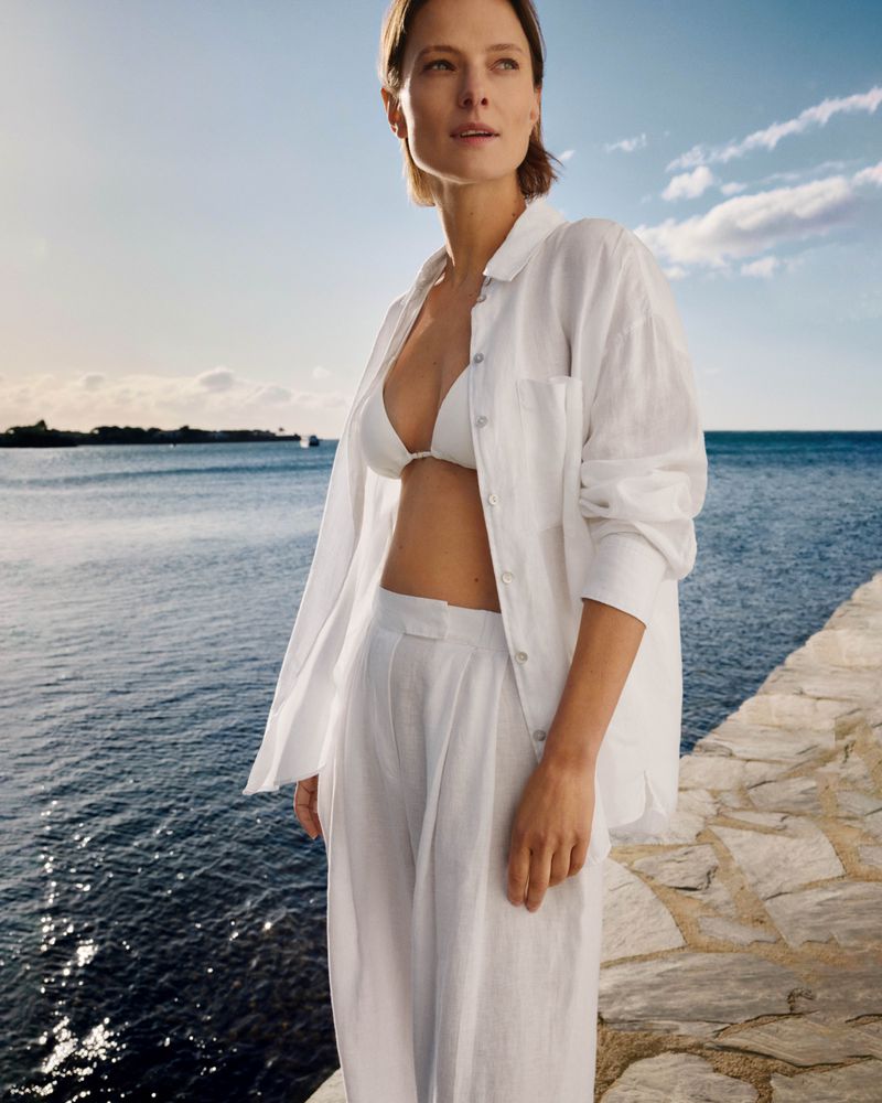 Adult woman in white linen shirt and pants stands on a stone path near the ocean with blue sky and clouds in a fashion shot.